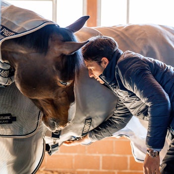 Steve Guerdat in a Horseware Signature jacket putting the Rambo Protector on his horse, in a stable.